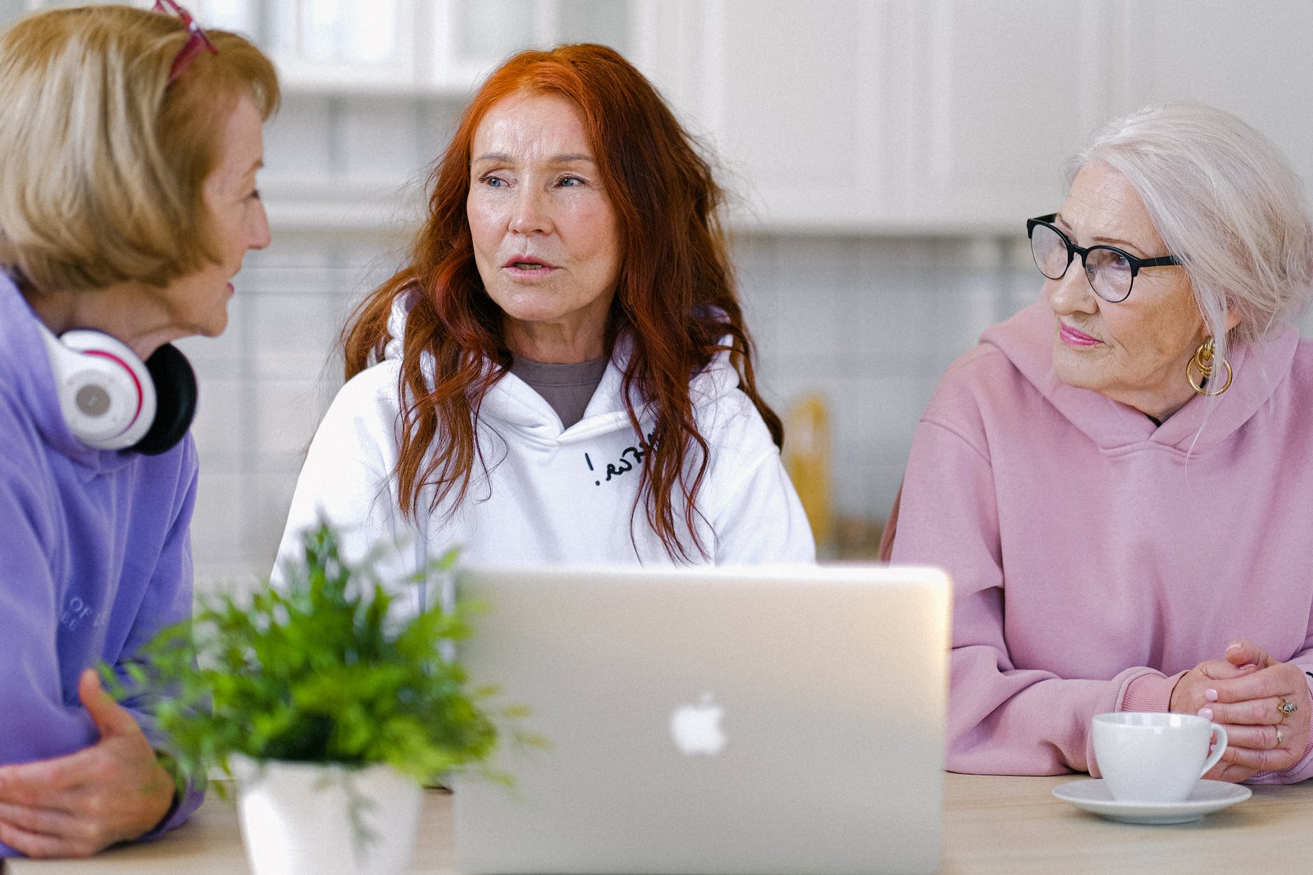 Three older women in casual clothes focused on each other as they have a discussion. A laptop, plant and teacup are on the counter in front of them.
