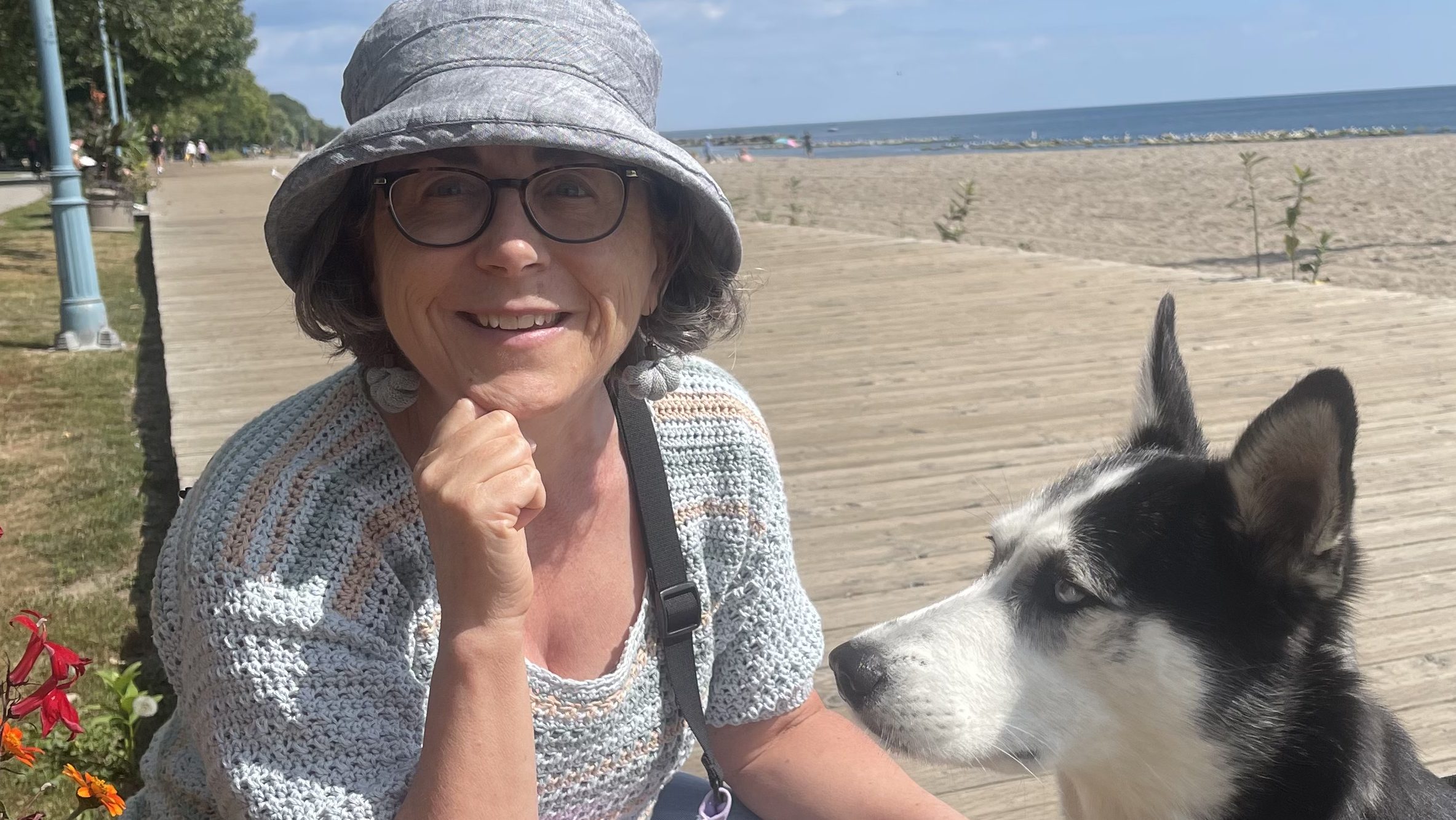 I am crouched down next to a husky and smiling. We are on the boardwalk and the beach is behind me. I am wearing a sunhat, glasses and a blue crocheted top.