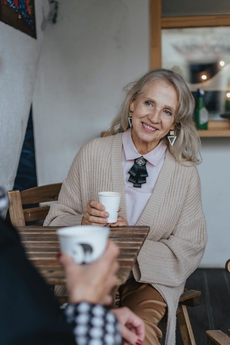 An older woman with hair and large geometric earrings hold a coffee cup and sits at a table smiling at the person who is sitting across from her.