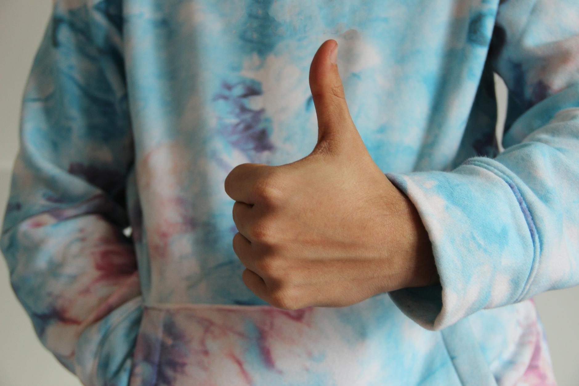 A person in a tie dye sweater is giving a thumbs up sign with one hand.